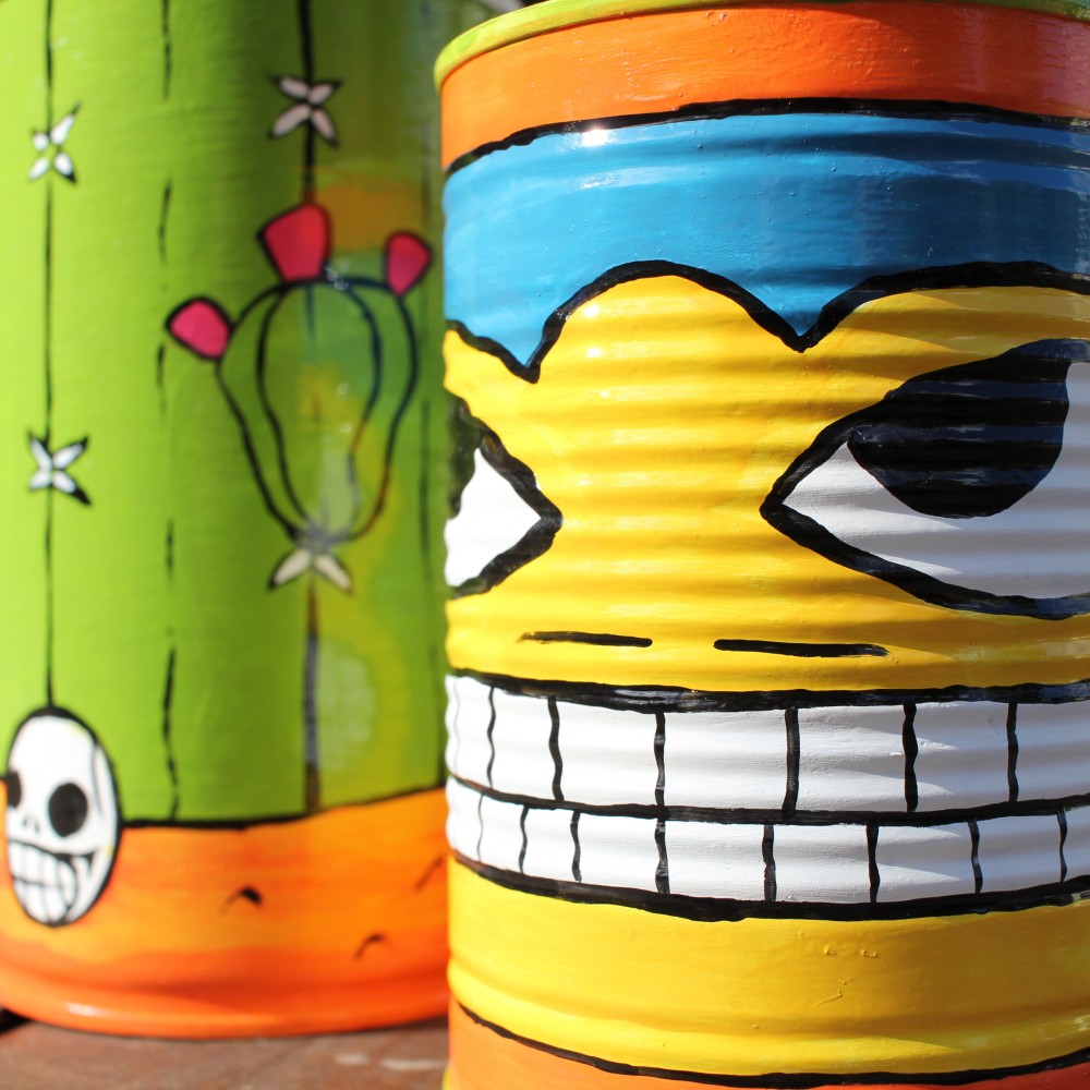 A phot of two tin cans painted in a mexican style using enamel paints. They are very bright. one resembles a mexican wrestler, the other resembles a cactus in the desert with a skull.