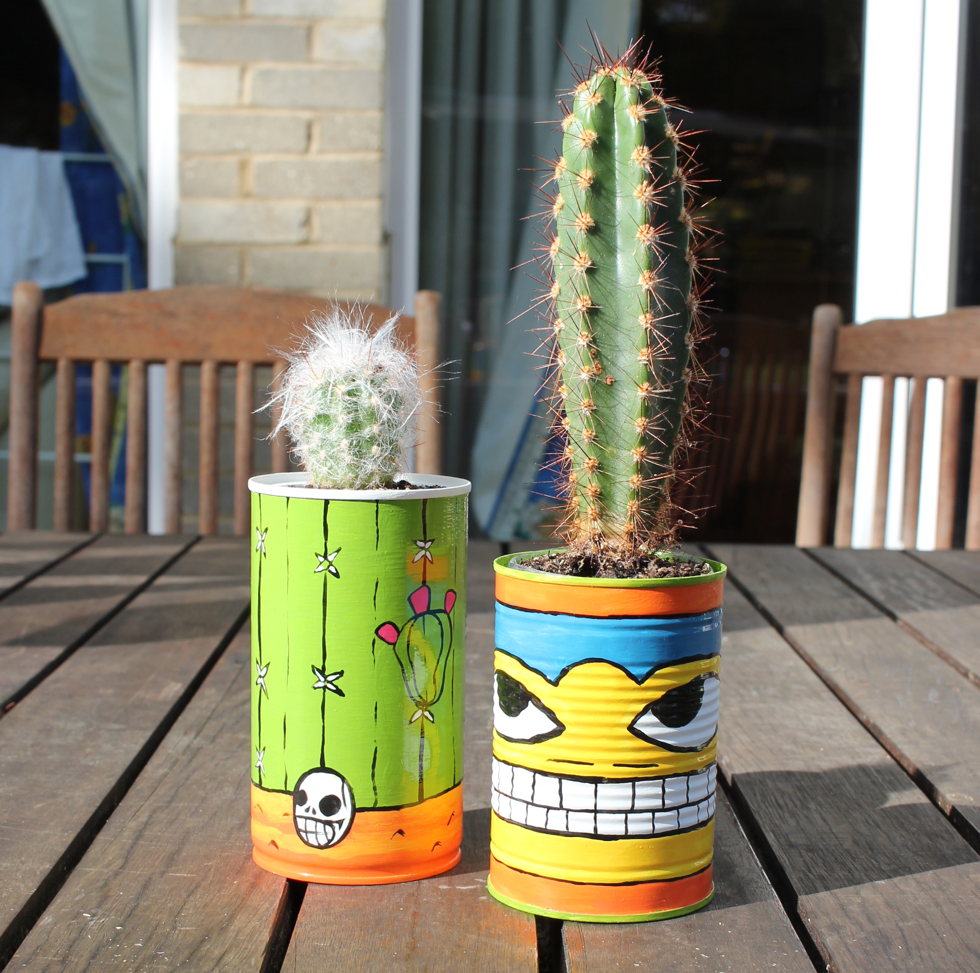 An image of two cactus pots with cacti. the pots are made from up-cycled tin cans. they are painted in a mexican art style in bright and vivid enamel paint.
